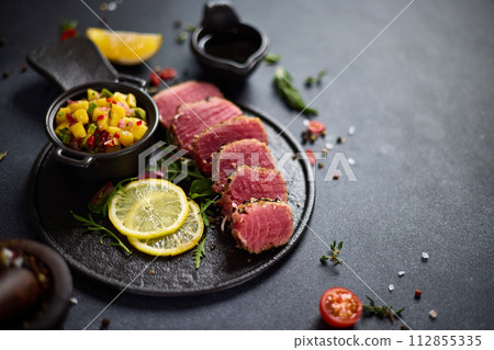 Sliced Organic grilled Tuna fillet covered with sesame seeds and mango salsa on black ceramic serving board 112855335