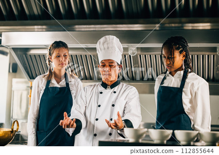 Professional Asian senior chef guides diverse students in a restaurant kitchen workshop focusing on teamwork learning and note-taking. Educational setting. Food Edocation 112855644