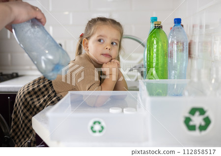 Little cute child helps mom sort plastic in the kitchen. Cute daughter puts plastic bottles in a box with a recycling logo. A mother teaches her child to sort garbage and take care of the planet. Little cute child helps mom sort plastic in the kitchen. Cute daughter puts plastic bottles in a box with a recycling logo. A mother teaches her child to sort garbage and take care of the planet. 112858517