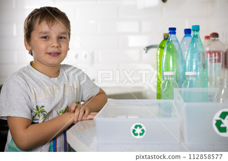 The child smiles, looking at the camera, ready to take apart the plastic. A child next to boxes with a recycling sign is ready to sort plastic. The concept of teaching a child to sort garbage. 112858577