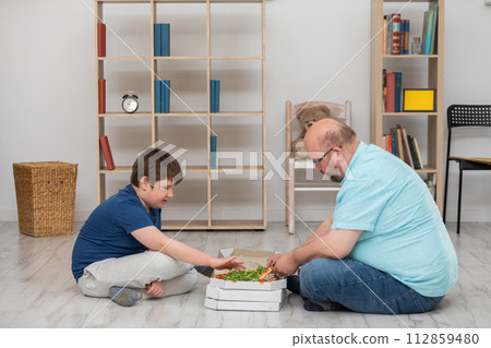Sitting on the floor, father and son pick out slices of pizza. Sitting on the floor, father and son pick out slices of pizza. 112859480