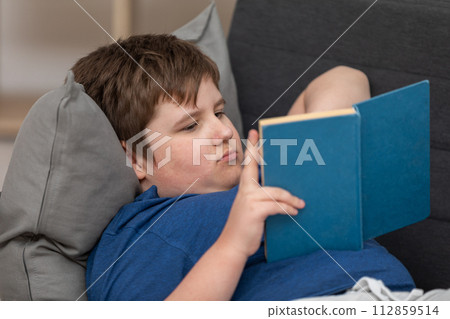A reclining young boy reads his school reading at home. 112859514