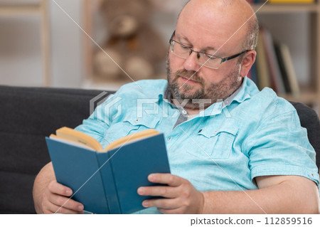 A bald man sitting on a sofa is reading a book at home. A bald man sitting on a sofa is reading a book at home. 112859516