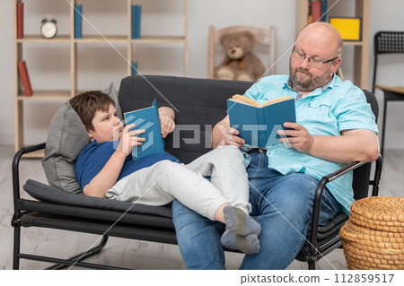 A father and son are reading books and sitting together on the sofa in the room. A father and son are reading books and sitting together on the sofa in the room. 112859517
