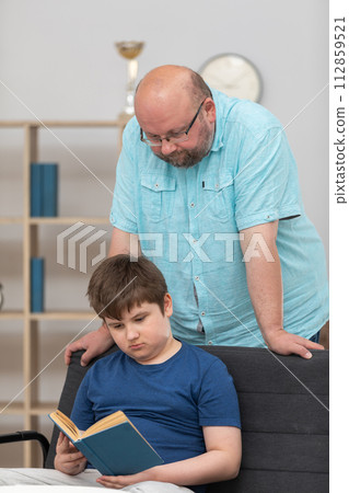 The son is reading a book and the father stands over him and checks. 112859521