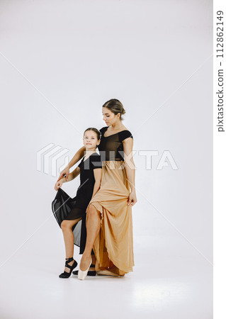 Little girl ballerina with her mother on white background. Brunette girl wearing black ballet suit and adult woman black and golden dress. Two female dancer dancing together in a pointe shoes. 112862149