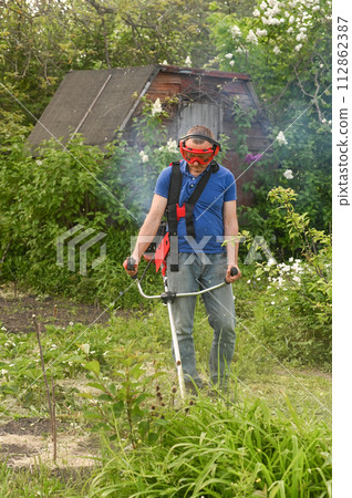 A worker mows the tall grass with a gasoline lawn mower in the garden A worker mows the tall grass with a gasoline lawn mower in the garden 112862387