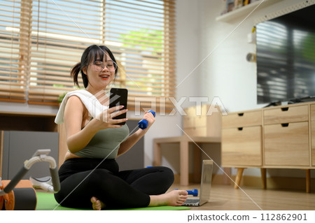 Smiling woman lifting dumbbell and using smartphone in living room. Home workout concept Smiling woman lifting dumbbell and using smartphone in living room. Home workout concept 112862901