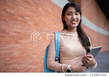 Young and cute Asian college student girls smile carrying a backpack with a textbooks in front of the university campus building. learning materials, importance of education becoming intelligent 112863159