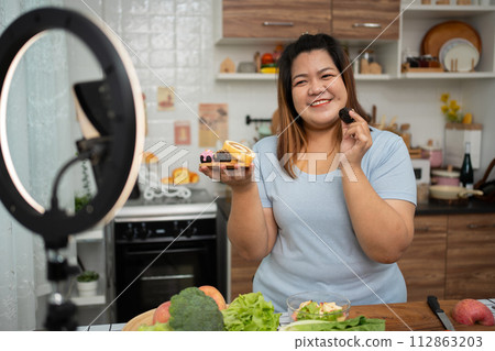Asian Pregnant blogger looking at smartphone camera, she is live streaming cooking class for pregnant. Asian woman standing happily smiling at the kitchen counter preparing fresh organic salad. Asian Pregnant blogger looking at smartphone camera, she is live streaming cooking class for pregnant. Asian woman standing happily smiling at the kitchen counter preparing fresh organic salad. 112863203