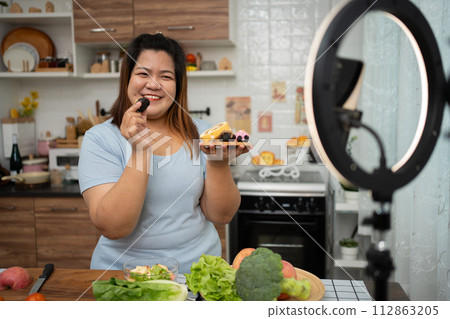Asian Pregnant blogger looking at smartphone camera, she is live streaming cooking class for pregnant. Asian woman standing happily smiling at the kitchen counter preparing fresh organic salad. 112863205