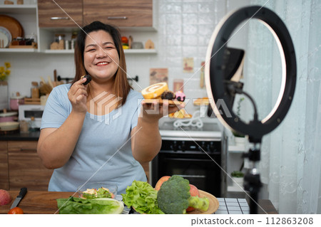 Asian Pregnant blogger looking at smartphone camera, she is live streaming cooking class for pregnant. Asian woman standing happily smiling at the kitchen counter preparing fresh organic salad. 112863208