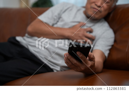 Senior male asian presses hand to his chest has a heart attack suffers from unbearable pain in the living room, lonely men try using the smartphone to call for Emergency help. Senior male asian presses hand to his chest has a heart attack suffers from unbearable pain in the living room, lonely men try using the smartphone to call for Emergency help. 112863381