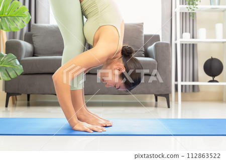 Close up of slim Asian woman in green top and leggings doing yoga in standing forward fold or Uttanasana on blue mat in living room at home in the morning.. 112863522