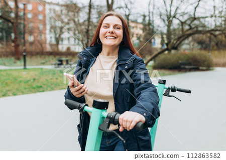 Smiling young woman standing near electric push scooter using smart phone. Ecological transport. Active lifestyle. Urban lifestyle concept. Smiling young woman standing near electric push scooter using smart phone. Ecological transport. Active lifestyle. Urban lifestyle concept. 112863582