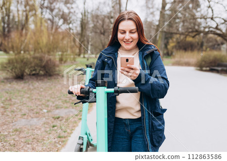 Smiling young woman standing near electric push scooter using smart phone. Ecological transport. Active lifestyle. Urban lifestyle concept. Smiling young woman standing near electric push scooter using smart phone. Ecological transport. Active lifestyle. Urban lifestyle concept. 112863586