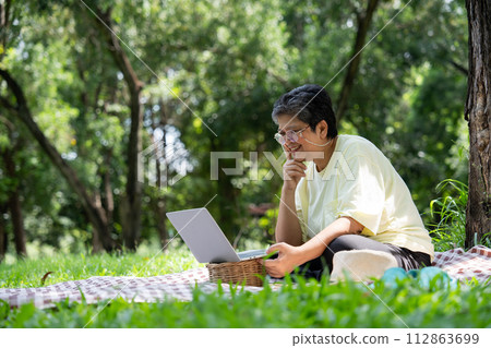 Happy Asian senior woman using laptop for working online outside office. Elderly retirement using laptop for learning new skill and freelance working in park, senior lifestyle with technology 112863699