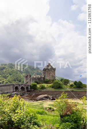 Historic Eilean Donan Castle, connected by stone bridge under cloudy sky, with fluttering flag Historic Eilean Donan Castle, connected by stone bridge under cloudy sky, with fluttering flag 112863768