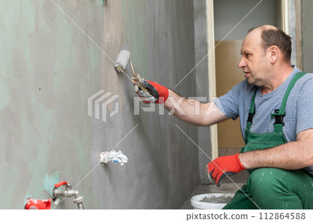 In the bathroom, a general construction worker applies moisture insulation using a roller. In the bathroom, a general construction worker applies moisture insulation using a roller. 112864588