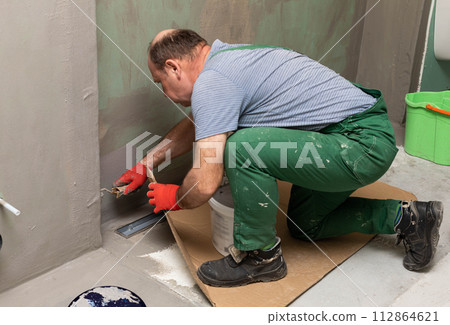 In the bathroom, a general construction worker applies moisture insulation using a roller. 112864621