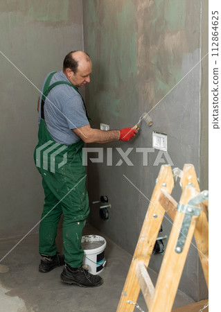 An experienced construction worker applies damp insulation with a paint roller to the wall. 112864625