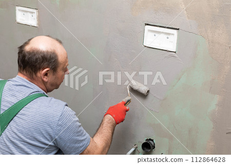 In the bathroom, a general construction worker applies moisture insulation using a roller. In the bathroom, a general construction worker applies moisture insulation using a roller. 112864628