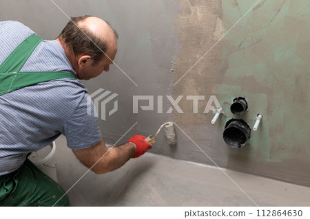 In the bathroom, a general construction worker applies moisture insulation using a roller. In the bathroom, a general construction worker applies moisture insulation using a roller. 112864630