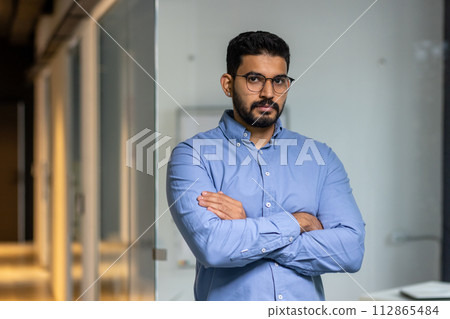 A man in a blue shirt is standing in front of a glass wall. He is wearing glasses and has his arms crossed. Concept of professionalism and confidence 112865484
