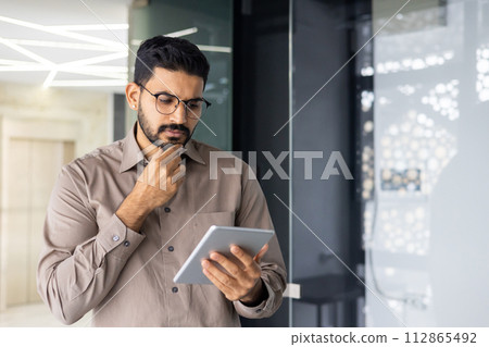 A man in a brown shirt is looking at a tablet. He is wearing glasses and he is deep in thought 112865492