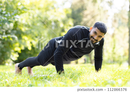 Active man in sportswear engaged in push-ups, showing strength and fitness at a sunny, green park. 112866376