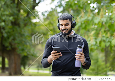 Smiling man in sportswear holding a water bottle while using his smartphone and wearing headphones in a park setting. 112866569