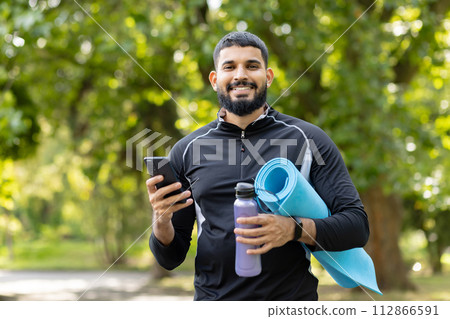Happy, fit man holding a yoga mat and water bottle while using his smartphone in a sunny, green park. Happy, fit man holding a yoga mat and water bottle while using his smartphone in a sunny, green park. 112866591