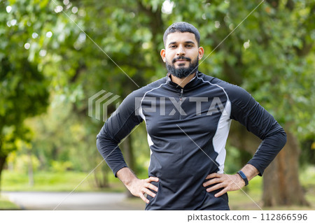 Focused athletic man with hands on hips standing outdoors, wearing sportswear in a park setting, exuding confidence and determination. Focused athletic man with hands on hips standing outdoors, wearing sportswear in a park setting, exuding confidence and determination. 112866596