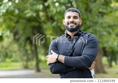 A fit man in sportswear stands with his arms crossed, exuding confidence amid the lush greenery of a serene park. A fit man in sportswear stands with his arms crossed, exuding confidence amid the lush greenery of a serene park. 112866599