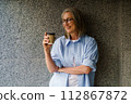 A woman wearing glasses stands holding a cup of coffee. 112867872
