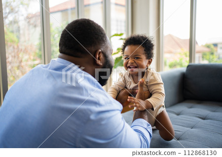Portrait Of Happy African American Dad With Cute Little Baby Girl on couch at home in the living room, caring father smiling and amusing his girl while sitting on the couch, happy family Portrait Of Happy African American Dad With Cute Little Baby Girl on couch at home in the living room, caring father smiling and amusing his girl while sitting on the couch, happy family 112868130