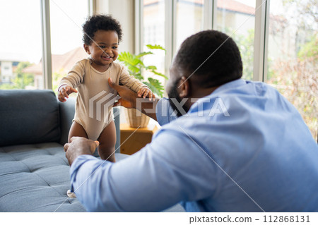 Portrait Of Happy African American Dad With Cute Little Baby Girl on couch at home in the living room, caring father smiling and amusing his girl while sitting on the couch, happy family Portrait Of Happy African American Dad With Cute Little Baby Girl on couch at home in the living room, caring father smiling and amusing his girl while sitting on the couch, happy family 112868131