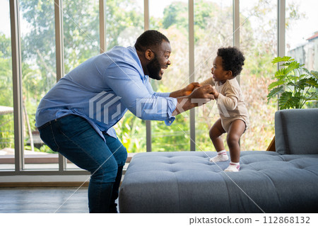 Portrait Of Happy African American Dad With Cute Little Baby Girl on couch at home in the living room, caring father smiling and amusing his girl while sitting on the couch, happy family 112868132