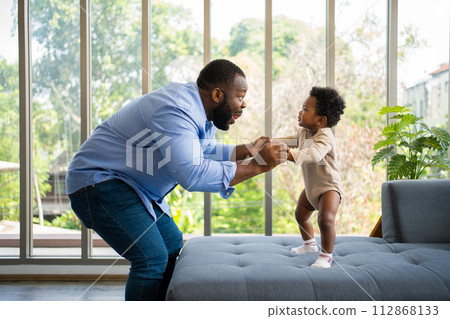 Portrait Of Happy African American Dad With Cute Little Baby Girl on couch at home in the living room, caring father smiling and amusing his girl while sitting on the couch, happy family Portrait Of Happy African American Dad With Cute Little Baby Girl on couch at home in the living room, caring father smiling and amusing his girl while sitting on the couch, happy family 112868133
