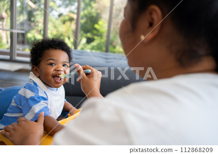 Asian mother feeding her 9 months old her cute little baby and African American helping for holding food plate At Home. Photo series of family, kids and happy people concept. Parents feed kids. Asian mother feeding her 9 months old her cute little baby and African American helping for holding food plate At Home. Photo series of family, kids and happy people concept. Parents feed kids. 112868209