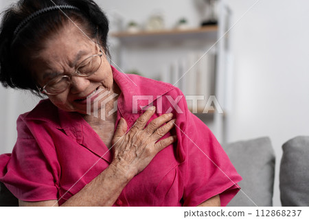 Asian senior elderly grandmother suffering from heart pain and holding her chest while sitting on the sofa in her living room. Medical emergency treatment and health care insurance protection concept Asian senior elderly grandmother suffering from heart pain and holding her chest while sitting on the sofa in her living room. Medical emergency treatment and health care insurance protection concept 112868237