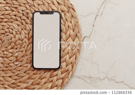 Smartphone with Blank screen on round rattan casket and beige marble background Smartphone with Blank screen on round rattan casket and beige marble background 112868336