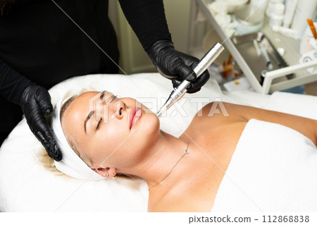 Microneedle RF lifting procedure. A cosmetologist performs a facial rejuvenation procedure on a woman. Hardware cosmetology. Microneedle RF lifting procedure. A cosmetologist performs a facial rejuvenation procedure on a woman. Hardware cosmetology. 112868838