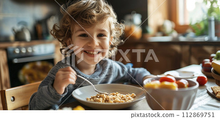 The child is having breakfast. The child eats cereal. Little boy at the dining table in the kitchen. Healthy balanced nutrition for children The child is having breakfast. The child eats cereal. Little boy at the dining table in the kitchen. Healthy balanced nutrition for children 112869751