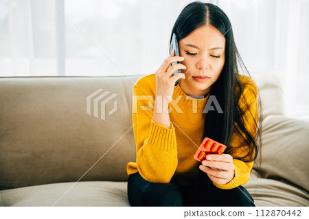 Highlighting consultation, Asian woman takes medicine consults doctor via phone while on couch at home. Depicting healthcare quarantine and social distancing amid coronavirus. 112870442