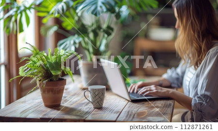 woman sitting at wooden table and working on laptop. A cup of coffee with a potted plant. The environment is bright and clean, a relaxed work environment 112871261