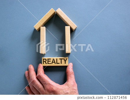 Male hand builds a model of a wooden house from wooden blocks. Word 'realty'. Copy space. Business concept. Beautiful blue background. 112871456