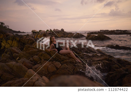 Woman reclines on coastal boulders at dusk, serene sea horizon, waves break nearby, tranquil vacation scene, sunset ocean view, solo traveler tranquility, female enjoying natures splendor. 112874422