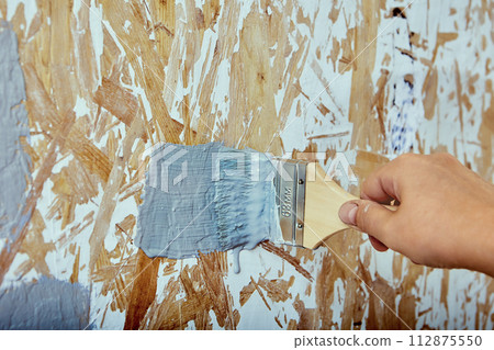Interior painting, house painter applies brush strokes with gray paint to chipboard wall. Interior painting, house painter applies brush strokes with gray paint to chipboard wall. 112875550