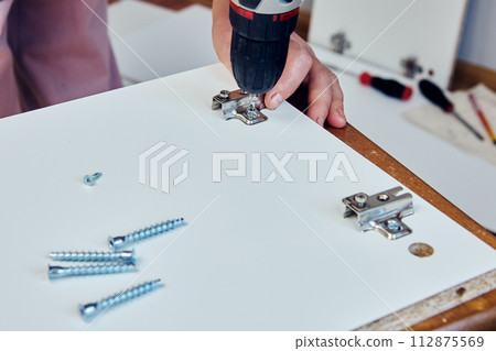 Assembly of flat pack furniture , adjustable hinges are installed in chipboard cabinet door. 112875569